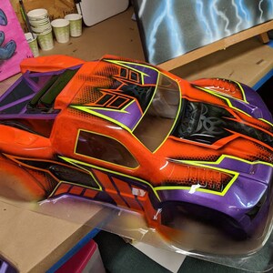 May include: A bright orange and purple painted body shell for a remote control truck. The shell has a black and yellow graphic design with a honeycomb pattern.