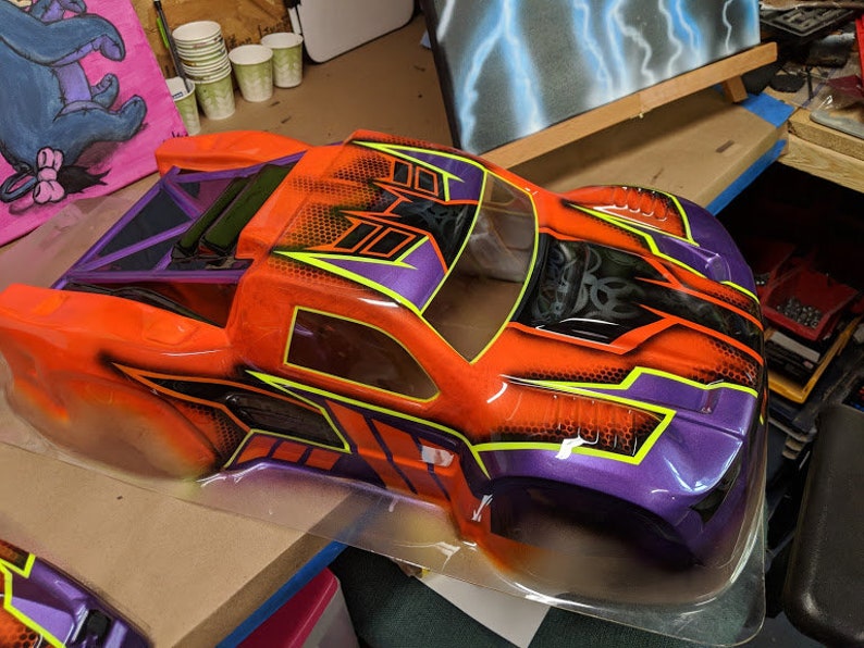 May include: A bright orange and purple painted body shell for a remote control truck. The shell has a black and yellow graphic design with a honeycomb pattern.