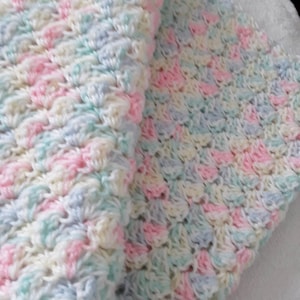 May include: A handmade crochet blanket in pastel colors. The blanket features a textured pattern with a combination of pink, blue, and cream-colored yarn. The soft, cozy blanket is folded, showcasing its intricate stitchwork and color variations.