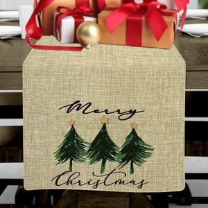 May include: A beige burlap table runner with the text "Merry Christmas" and three green Christmas trees with gold stars on top. The runner is on a wooden table with wrapped presents and gold ornaments.