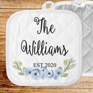 May include: White quilted pot holders with a floral design and the text "The Williams EST 2020". The pot holders have a loop for hanging and a diamond pattern. The floral design features blue flowers and green leaves.