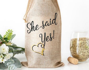 May include: A burlap wine bottle bag with a gold heart and the text "She said Yes!" printed on it. The bag is sitting on a white surface with a glass of champagne and a cork.