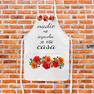 May include: White kitchen apron with a floral design and the text "nadie me ayuda en esta casa" in black.