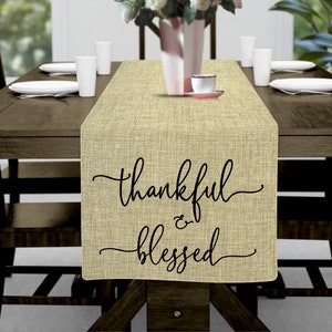 May include: A beige burlap table runner with the text "thankful & blessed" in black cursive script. The runner is on a wooden table with two chairs and a white cup and saucer.