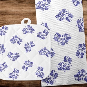 May include: A white quilted pot holder and a matching kitchen towel, both decorated with a repeating pattern of blue floral designs. The pot holder has a hanging loop. The towel has a textured surface.