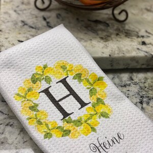 May include: White kitchen towel with a waffle weave texture. The towel features a yellow lemon wreath design around a large black letter "H". The name "Heine" is printed below the letter, with "EST 1981" below the name.