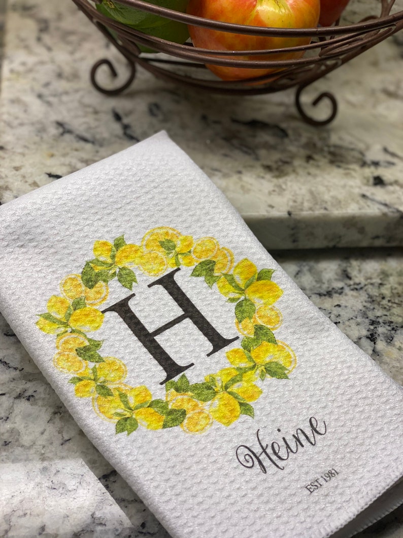 May include: White kitchen towel with a waffle weave texture. The towel features a yellow lemon wreath design around a large black letter "H". The name "Heine" is printed below the letter, with "EST 1981" below the name.