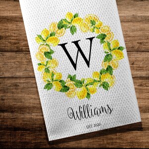 May include: White kitchen towel with a lemon wreath design. The wreath surrounds a large black letter "W". The word "Williams" is written in cursive below the letter, with "EST 2020" underneath. The towel is on a wooden surface.
