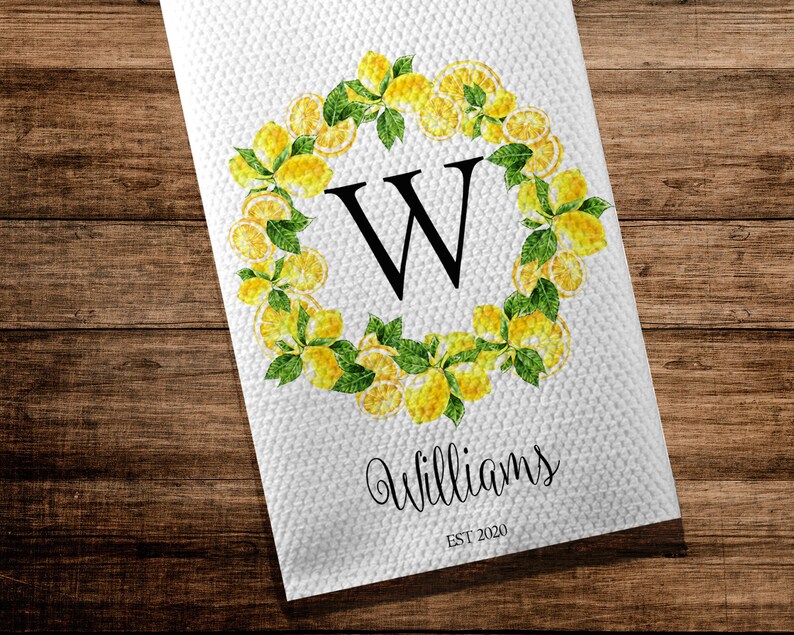 May include: White kitchen towel with a lemon wreath design. The wreath surrounds a large black letter "W". The word "Williams" is written in cursive below the letter, with "EST 2020" underneath. The towel is on a wooden surface.