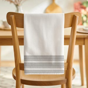 May include: A white waffle-weave kitchen towel with a decorative gray border hangs on a wooden chair. The towel's texture is visible, and the border features a repeating pattern of lines and curves.