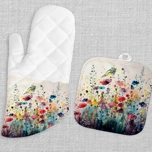 May include: White oven mitt and pot holder set. The set features a watercolor design of colorful wildflowers and a small bird. The mitt has a quilted pattern, and the pot holder has a hanging loop. The design includes red, blue, and yellow flowers.