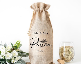 May include: A beige burlap wine bottle bag with a drawstring closure. The bag is printed with "Mr. & Mrs. Patton est. 2021" in black script. A wine glass with gold glitter and a cork are next to the bag.
