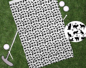 Może przedstawiać: Biały ręcznik golfowy z czarnym wzorem sylwetki kotów. Ręcznik leży na zielonej trawie z dwiema białymi piłkami golfowymi i kijem golfowym.