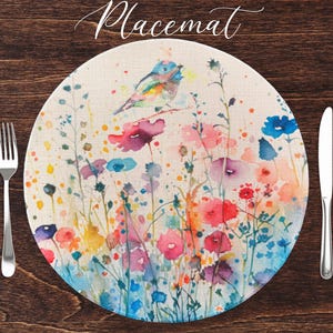 May include: Round placemat featuring a watercolour design of a bird and flowers. The placemat has a linen texture and is beige with splashes of pink, blue, yellow, and green.