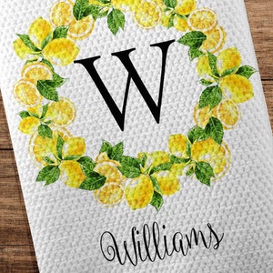 May include: White kitchen towel with a lemon wreath design. The wreath surrounds a large black letter "W" and is made of yellow lemons and green leaves. The name "Williams" is written below the letter, with "EST 2020" underneath.