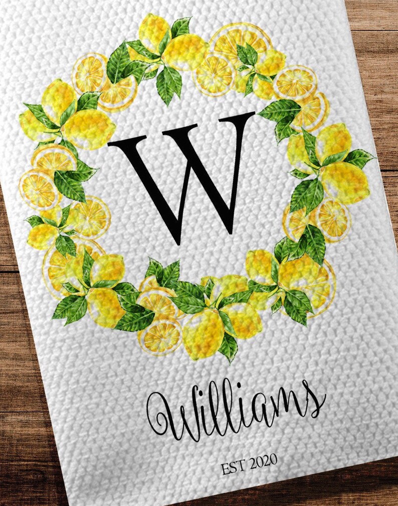 May include: White kitchen towel with a lemon wreath design. The wreath surrounds a large black letter "W" and is made of yellow lemons and green leaves. The name "Williams" is written below the letter, with "EST 2020" underneath.