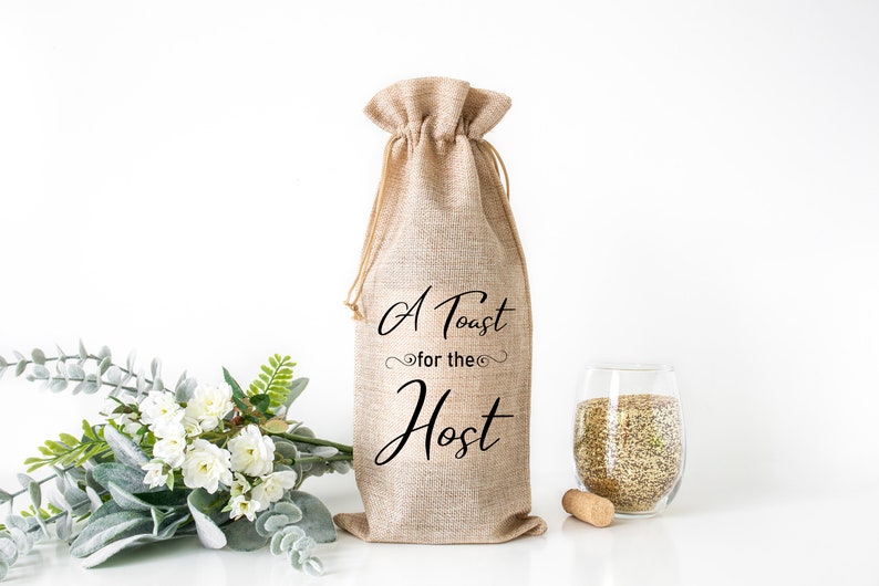 Może przedstawiać: Brązowy worek z juty na butelkę wina z czarnym napisem "A Toast for the Host".