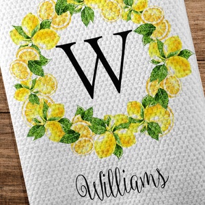 May include: White kitchen towel with a lemon wreath design. The wreath surrounds a large black letter "W" and is made of yellow lemons and green leaves. The name "Williams" and the year "EST 2020" are printed below the wreath.