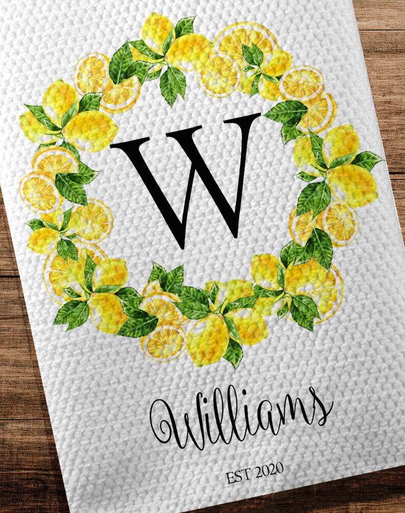 May include: White kitchen towel with a lemon wreath design. The wreath surrounds a large black letter "W" and is made of yellow lemons and green leaves. The name "Williams" and the year "EST 2020" are printed below the wreath.