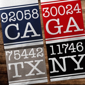 May include: Four rectangular fabric panels with white numbers and state abbreviations. The top left panel is navy blue with white text "92058 CA". The top right panel is red with white text "30024 GA". The bottom left panel is grey with white text "75442 TX". The bottom right panel is black with white text "11746 NY".