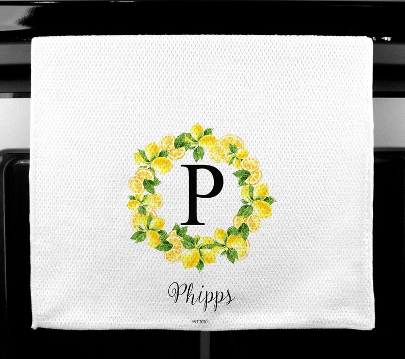 May include: White kitchen towel with a lemon wreath design. The wreath surrounds a large black letter "P". The name "Phipps" is written in cursive below the wreath, with "EST 2020" below the name.