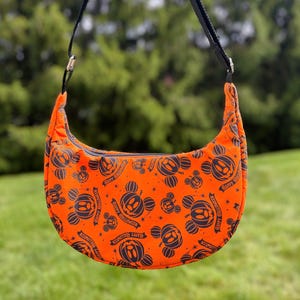 Puede incluir: Bolso de hombro naranja en forma de media luna con una correa negra. El bolso está estampado con cabezas de Mickey Mouse con forma de calabaza negras y las palabras "Happy Halloween". El bolso tiene una cremallera negra y se sostiene sobre un fondo verde.