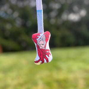 K&ouml;nnte beinhalten: Ein wei&szlig;er Schn&uuml;rsenkel mit einem blauen Abschnitt und einem kleinen Anh&auml;nger mit einer Cartoonfigur in roter Kleidung. Der Anh&auml;nger hat einen grimmigen Gesichtsausdruck.