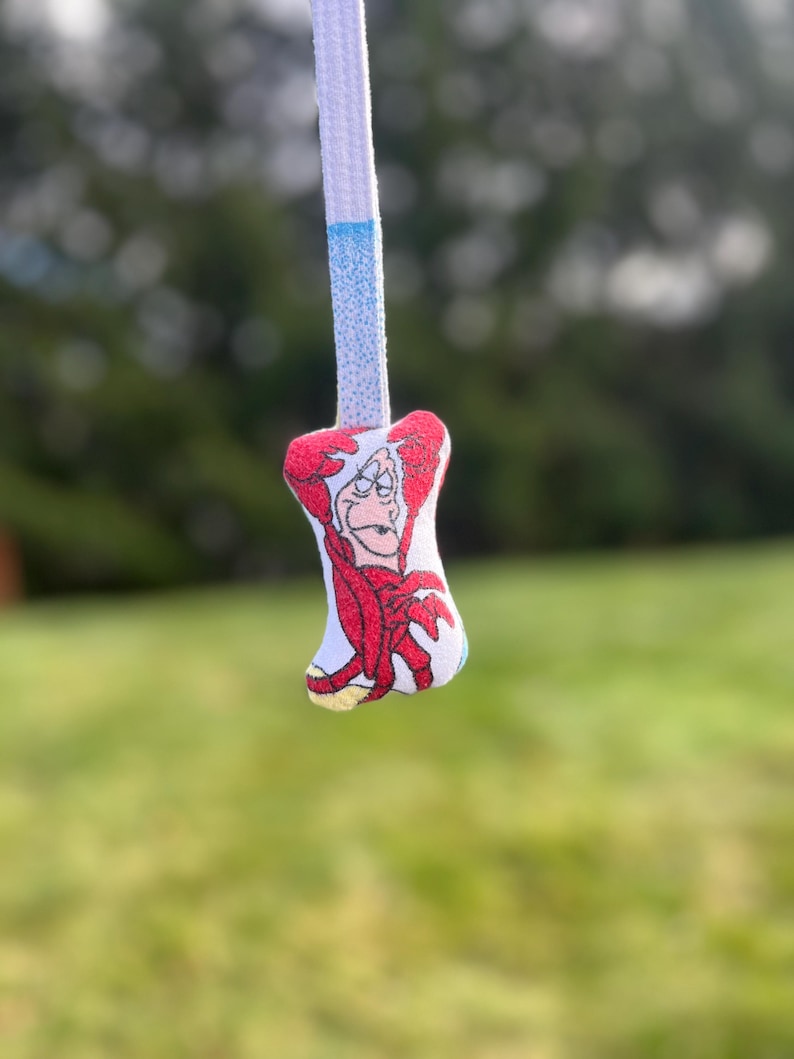 K&ouml;nnte beinhalten: Ein wei&szlig;er Schn&uuml;rsenkel mit einem blauen Abschnitt und einem kleinen Anh&auml;nger mit einer Cartoonfigur in roter Kleidung. Der Anh&auml;nger hat einen grimmigen Gesichtsausdruck.