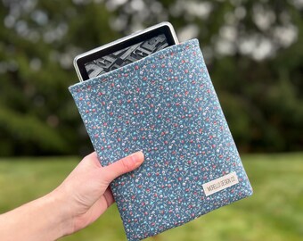 Op de afbeelding: Een blauwe e-reader hoes met bloemenprint en een wit label met de tekst "MORELLO DESIGN CO.". De hoes bevat een e-reader. De achtergrond is een vage groene gras en bomen.
