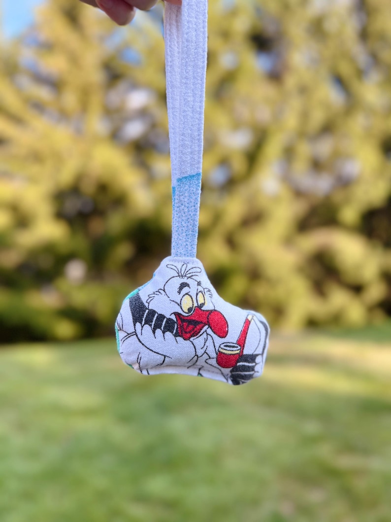 Puede incluir: Un juguete de peluche con un personaje de dibujos animados con una nariz roja y una pipa, sobre un fondo verde borroso. El juguete es blanco con detalles en negro y rojo, y una correa blanca.