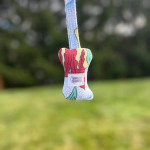 K&ouml;nnte beinhalten: Ein farbenfroher Gitarrenplektrumhalter aus Stoff mit passendem Gurt. Der Plektrumhalter hat ein lebendiges Design mit roten, gelben und gr&uuml;nen Akzenten. Auf dem Gurt und dem Plektrumhalter steht der Text "MORELLO DESIGN CO."