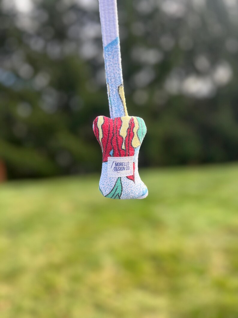 K&ouml;nnte beinhalten: Ein farbenfroher Gitarrenplektrumhalter aus Stoff mit passendem Gurt. Der Plektrumhalter hat ein lebendiges Design mit roten, gelben und gr&uuml;nen Akzenten. Auf dem Gurt und dem Plektrumhalter steht der Text "MORELLO DESIGN CO."