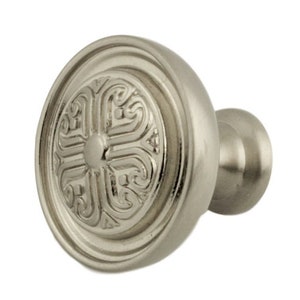 May include: A silver cabinet knob with a decorative, ornate design. The knob has a round base and a raised, circular center.
