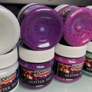 May include: A collection of small jars of glitter paste. The jars are white with white lids and labels that read "Pentart Glitter Paste." The glitter paste colors include white, purple, pink, and green. The jars are stacked on top of each other.