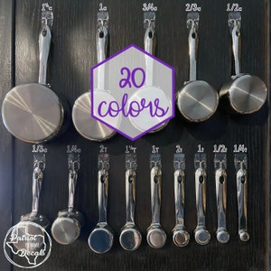 May include: A set of 12 stainless steel measuring cups hanging on a black wall. The cups are labeled with measurements in cups and tablespoons. The text "20 colors" is written in purple on a white hexagon.