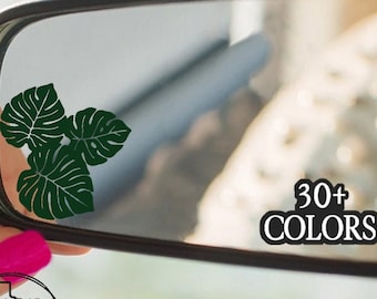 Può includere: Uno specchietto retrovisore con un adesivo a forma di foglia di monstera verde. L'adesivo ha la forma di tre foglie. Il testo "30+ COLORS" è visibile nel riflesso dello specchio.