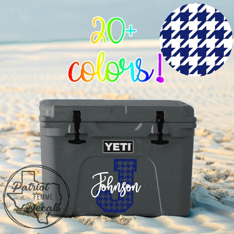 Op de afbeelding: Een grijze YETI-koeler met zwarte grendels en de tekst "YETI" op de voorkant. Een blauw en wit hondenpoot "U" staat op de voorkant, met de naam "Johnson" in witte letters. De tekst "20+ kleuren!" is in regenboogkleuren.