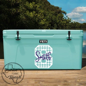 Op de afbeelding: Een lichtblauwe Yeti-koeler met een wit en blauw hondenpoottandnummer 8 en de naam "Smith" in een scriptlettertype. De koeler heeft zwarte grendels en een zwart Yeti-logo. De koeler staat op een houten steiger met een meer en bomen op de achtergrond.