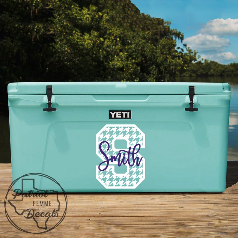 Op de afbeelding: Een lichtblauwe Yeti-koeler met een wit en blauw hondenpoottandnummer 8 en de naam "Smith" in een scriptlettertype. De koeler heeft zwarte grendels en een zwart Yeti-logo. De koeler staat op een houten steiger met een meer en bomen op de achtergrond.