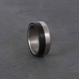 Op de afbeelding: Een close-up van een tweekleurige ring. De ring heeft een brede band van zwart materiaal en een gepolijst zilverkleurig metaal. De ring staat tegen een grijze achtergrond. De ring heeft een modern design.