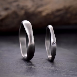Könnte beinhalten: Zwei silberne Eheringe. Einer ist schlicht, der andere mit einem kleinen Diamanten besetzt. Die Ringe haben eine glatte, matte Oberfläche und ein einzigartiges, geschwungenes Design. Sie werden auf einer dunklen, strukturierten Oberfläche präsentiert.