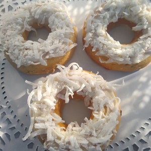 Puede incluir: Tres donuts con glaseado blanco y coco rallado en la parte superior. Los donuts están sobre un mantel de encaje blanco.