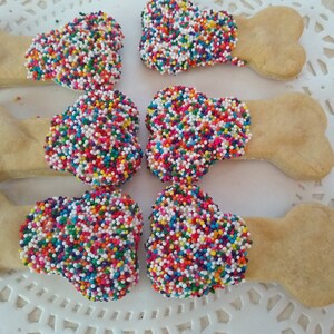 Puede incluir: Seis galletas con forma de hueso para perros decoradas con chispas de colores. Las galletas están dispuestas sobre un encaje blanco.