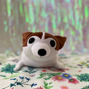 Puede incluir: Un juguete de peluche de perro blanco y marrón con ojos negros y una nariz negra. El juguete está sentado en una tela blanca con un patrón floral.