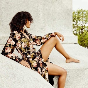 Peut inclure: Une femme portant une robe à manches longues noire à imprimé floral est assise sur un rebord de pierre. Elle a les cheveux bouclés et porte des boucles d'oreilles.
