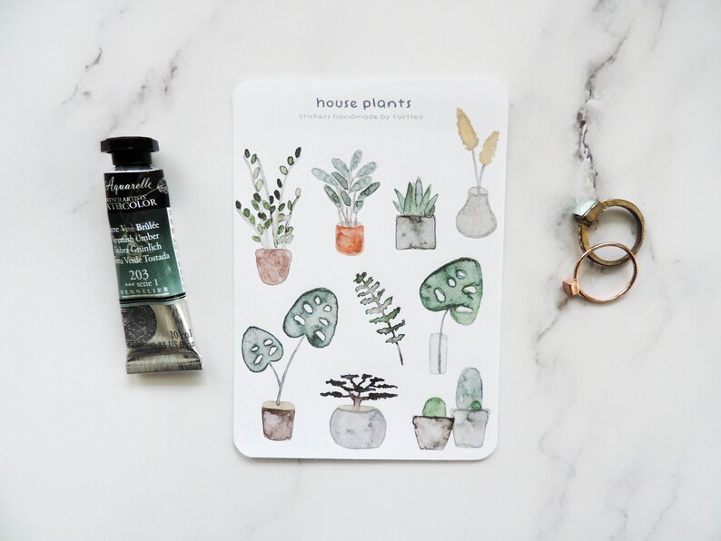 May include: A sheet of stickers featuring illustrations of house plants in pots. The stickers are watercolor style and feature a variety of plants, including ferns, succulents, and monstera leaves. The text "house plants" and "stickers handmade by turtle" is printed on the sheet.
