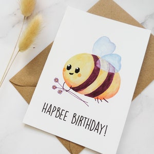 May include: A white card with a watercolour illustration of a cartoon bee holding flowers. The text "Hapbee Birthday!" is written in black below the bee.