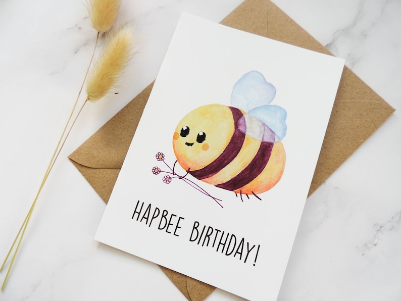 May include: A white card with a watercolour illustration of a cartoon bee holding flowers. The text "Hapbee Birthday!" is written in black below the bee.