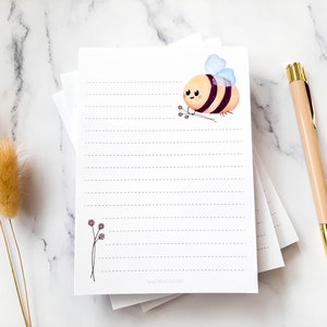 Puede incluir: Una pila de papel blanco rayado con una linda ilustración de abeja de dibujos animados. La abeja es amarilla y negra con un ala azul claro. La abeja sostiene un pequeño ramo de flores. El papel tiene una línea punteada para escribir.