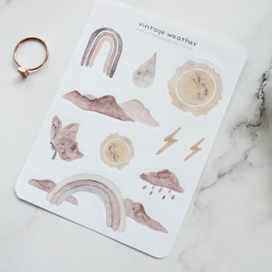 Puede incluir: Una hoja de pegatinas con tem&aacute;tica de clima vintage con ilustraciones de arco&iacute;ris, nubes, rayos, mariposas y soles en estilo acuarela. Las pegatinas est&aacute;n sobre un fondo blanco y tienen el texto "vintage weather" impreso en la parte superior.
